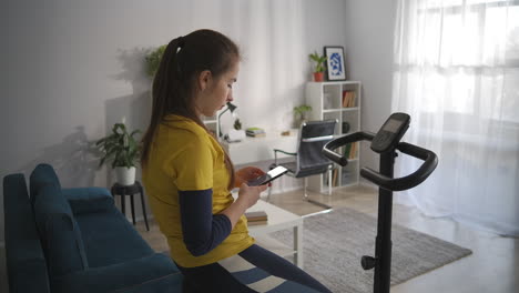 teen-girl-is-reading-messages-and-news-in-social-media-by-cell-sitting-on-stationary-bicycle-in-home-sport-workout-for-helath-and-internet-addiction